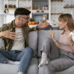 Man and woman arguing on a gray sofa in a modern living room with a kitchen in the background, mid-conversation.