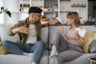 Man and woman arguing on a gray sofa in a modern living room with a kitchen in the background, mid-conversation.