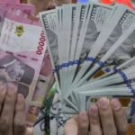 Hands hold a fan of cash: pink Thai baht notes on the left and fanned US $100 bills on the right.