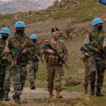 Group of soldiers in camouflage uniforms, some wearing blue UN helmets, marching with rifles along a rocky hillside trail.