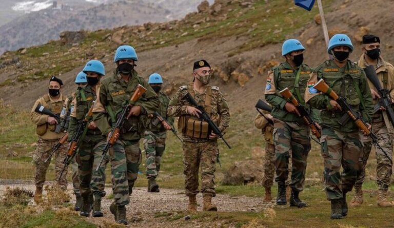 Group of soldiers in camouflage uniforms, some wearing blue UN helmets, marching with rifles along a rocky hillside trail.