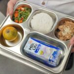Person holds a metal cafeteria tray with four compartments: mixed vegetables, white rice, shredded chicken with sesame seeds, and a peeled orange; plus a milk carton on the tray.