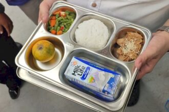 Person holds a metal cafeteria tray with four compartments: mixed vegetables, white rice, shredded chicken with sesame seeds, and a peeled orange; plus a milk carton on the tray.