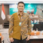 Smiling man in a batik shirt with a lanyard badge stands in a conference room with an Indonesian flag and stage backdrop behind him.