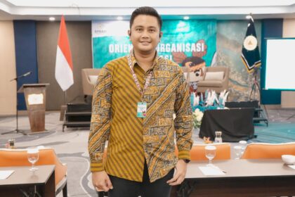 Smiling man in a batik shirt with a lanyard badge stands in a conference room with an Indonesian flag and stage backdrop behind him.