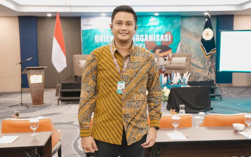 Smiling man in a batik shirt with a lanyard badge stands in a conference room with an Indonesian flag and stage backdrop behind him.
