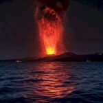 Nighttime volcanic eruption with lava plume shooting above a dark coastline, lava reflected on the water surface
