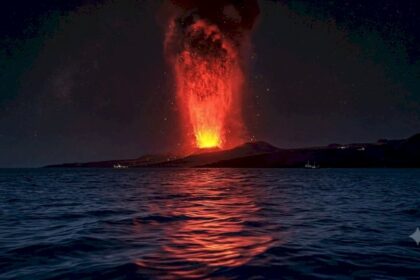 Nighttime volcanic eruption with lava plume shooting above a dark coastline, lava reflected on the water surface