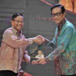 Two men on a stage shake hands while one presents a plaque featuring a portrait, during a formal event.