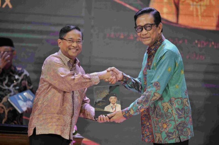 Two men on a stage shake hands while one presents a plaque featuring a portrait, during a formal event.