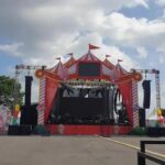 Outdoor carnival stage with a bright pink circus tent, large video screens, and speakers, set up for a performance on a sunny day.