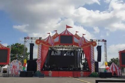 Outdoor carnival stage with a bright pink circus tent, large video screens, and speakers, set up for a performance on a sunny day.