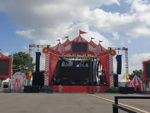 Outdoor carnival stage with a bright pink circus tent, large video screens, and speakers, set up for a performance on a sunny day.