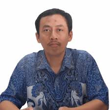 Man in a blue patterned shirt seated at a table, looking at the camera against a white background.