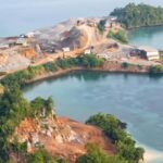 Aerial view of a small rocky island with exposed dirt and excavation equipment along winding paths, surrounded by calm blue water.