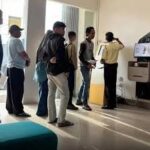 People lined up indoors at a service desk with a self-service kiosk on the right, holding documents while staff assist at the counter.