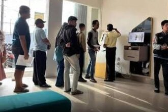 People lined up indoors at a service desk with a self-service kiosk on the right, holding documents while staff assist at the counter.