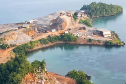 Aerial view of a small rocky island with exposed dirt and excavation equipment along winding paths, surrounded by calm blue water.