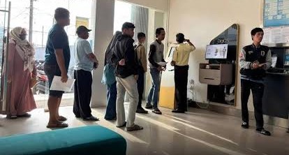 People lined up indoors at a service desk with a self-service kiosk on the right, holding documents while staff assist at the counter.
