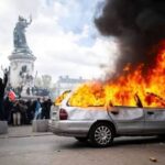 Silver sedan engulfed in flames on a city street as protesters stand nearby with a statue in the background.