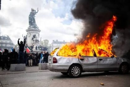 Silver sedan engulfed in flames on a city street as protesters stand nearby with a statue in the background.