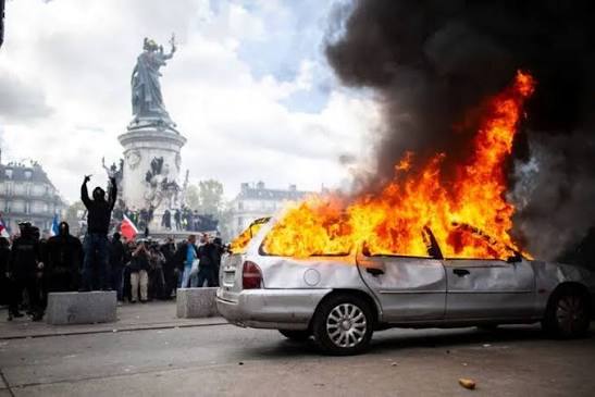Silver sedan engulfed in flames on a city street as protesters stand nearby with a statue in the background.