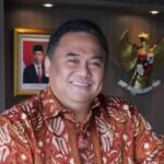 Smiling man in batik shirt seated in an office with an Indonesian Garuda emblem on the wall to his right.