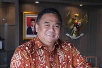 Smiling man in batik shirt seated in an office with an Indonesian Garuda emblem on the wall to his right.