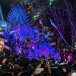 Colorful parade float with blue-purple feathered dragon shape surrounded by a crowd at night, during a festival show.