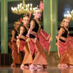 Four female dancers in gold and black traditional outfits perform on a stage with chandeliers.] - Note: remove extra bracket.