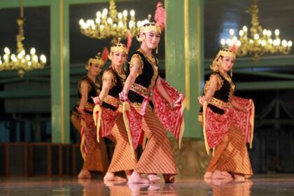 Four female dancers in gold and black traditional outfits perform on a stage with chandeliers.] - Note: remove extra bracket.