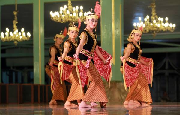 Four female dancers in gold and black traditional outfits perform on a stage with chandeliers.] - Note: remove extra bracket.