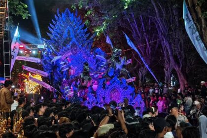 Colorful parade float with blue-purple feathered dragon shape surrounded by a crowd at night, during a festival show.