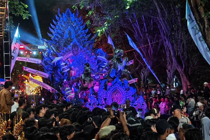 Colorful parade float with blue-purple feathered dragon shape surrounded by a crowd at night, during a festival show.