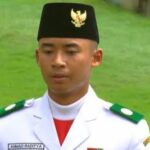 Young man in white ceremonial military uniform with green epaulettes and a black cap, standing on grass; name tag reads Ahmad Raditya.