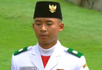 Young man in white ceremonial military uniform with green epaulettes and a black cap, standing on grass; name tag reads Ahmad Raditya.