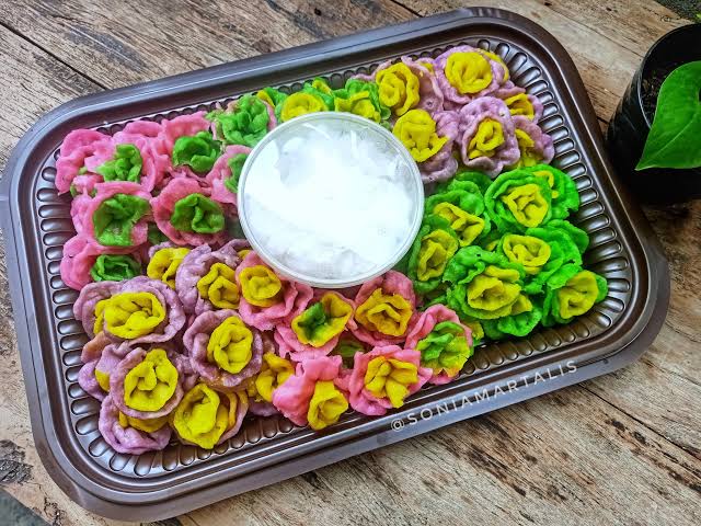 Colorful flower-shaped dumplings in pink, purple, yellow, and green arranged on a brown tray with a bowl of ice in the center.