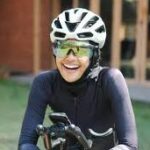 Cyclist wearing a white helmet and reflective sunglasses, smiling while holding handlebars on a bike.