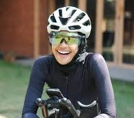 Cyclist wearing a white helmet and reflective sunglasses, smiling while holding handlebars on a bike.