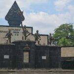 War memorial with four bronze soldier statues on a dark stone pedestal, central emblem, and plaques, set against a blue sky and palm trees.
