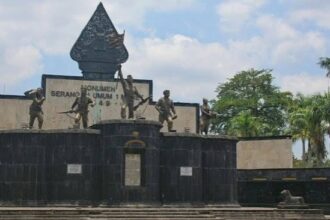 War memorial with four bronze soldier statues on a dark stone pedestal, central emblem, and plaques, set against a blue sky and palm trees.