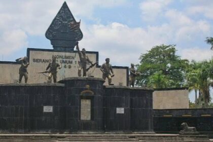 War memorial with four bronze soldier statues on a dark stone pedestal, central emblem, and plaques, set against a blue sky and palm trees.