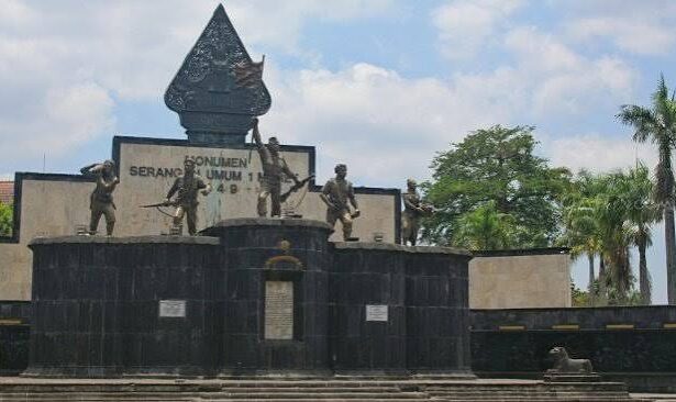 War memorial with four bronze soldier statues on a dark stone pedestal, central emblem, and plaques, set against a blue sky and palm trees.