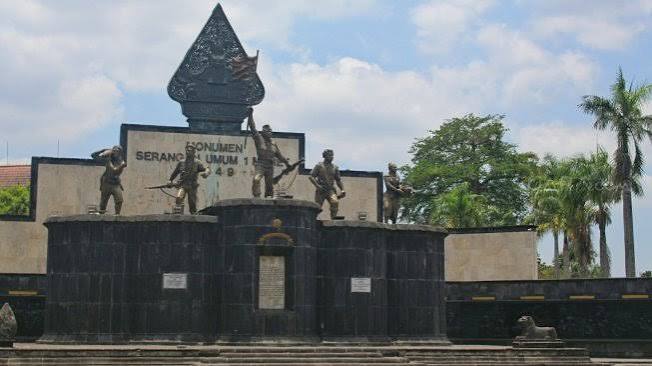 War memorial with four bronze soldier statues on a dark stone pedestal, central emblem, and plaques, set against a blue sky and palm trees.
