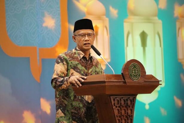 Man wearing batik shirt and black cap speaks at a wooden podium with a microphone on a colorful backdrop.