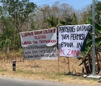 Two handwritten protest banners beside a road, criticizing local officials' integrity and permit handling, in a dry rural area.