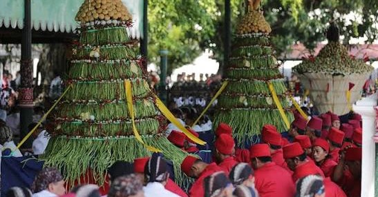 Ceremonial towers made from palm leaves and nuts with yellow ribbons, surrounded by people in red outfits at a festival.