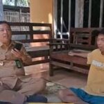 Two men sit on the floor and chat on a veranda; one wears a khaki uniform and holds a phone, the other wears a yellow T-shirt and blue shorts.