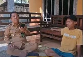 Two men sit on the floor and chat on a veranda; one wears a khaki uniform and holds a phone, the other wears a yellow T-shirt and blue shorts.