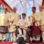 Group of men in cream traditional outfits standing on a decorated stage, with a small dog at their feet in front.